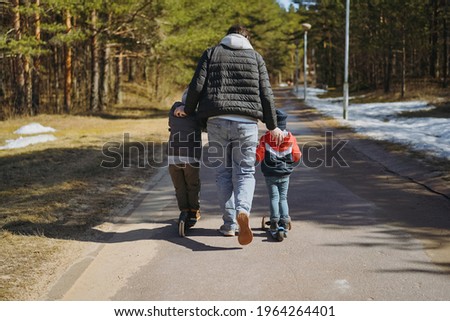 young man wearing hoodie, puffer jacket and  jeans pushing his two sons riding kick scooters along alley in park. Image with selective focus