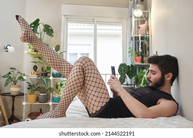 Young man wearing make up, fishnet stockings and lingerie using the smartphone laying on bed. Transgender.