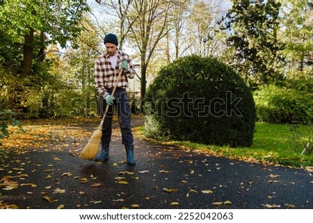 Young man wearing plaid shirt sweeping autumn leaves in park