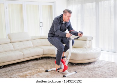 Young Man Wearing Sportswear While Exercising On A Spin Bike In The Living Room. Shot At Home