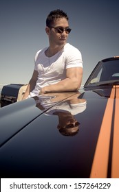 Young Man Wearing Tight White And Sunglasses Leaning Against Muscle Car With Racing Stripes