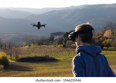 Young man wearing a VR glasses is piloting an fpv drone, he is sitting in a field at sunset. Image with toning and noise. Selective focus on drone