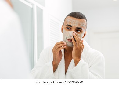 Young man  wearing white bathrobe applying facial mask in bathroom