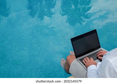 Young Man Wearing White Linen Shirt Working On Laptop By The Pool, Dipping His Feet In A Water. Male Travel Blogger Typing On Keyboard. Perks Of Being A Freelancer. Close Up, Copy Space, Background.