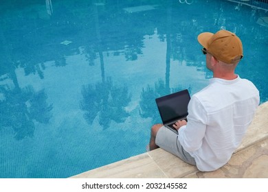 Young Man Wearing White Linen Shirt Working On Laptop By The Pool, Dipping His Feet In A Water. Male Travel Blogger Typing On Keyboard. Perks Of Being A Freelancer. Close Up, Copy Space, Background.