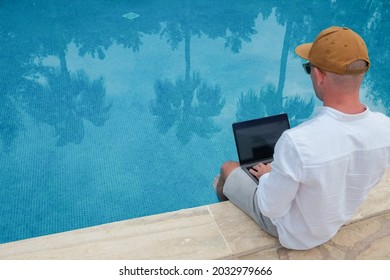 Young Man Wearing White Linen Shirt Working On Laptop By The Pool, Dipping His Feet In A Water. Male Travel Blogger Typing On Keyboard. Perks Of Being A Freelancer. Close Up, Copy Space, Background.