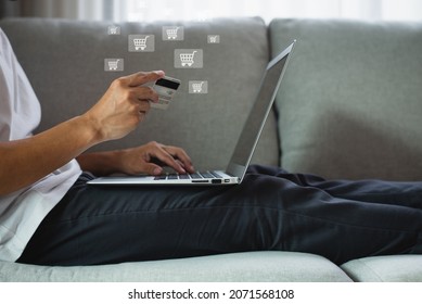 Young Man Wearing White T-shirt Sitting On Sofa In Living Room Using Laptop And Smartphone And Hands Holding Credit Card With Using Shopping Cart Icon. Show That He Is Shopping Online.