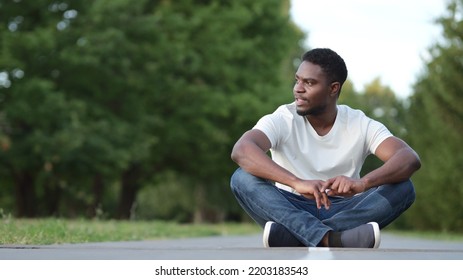 Young Man Wearing White T-shirt And Jeans Looks Around Sitting Cross Legged On Asphalt Against Green Trees. African American Guy Enjoys Resting In City Park