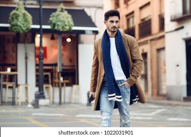Young man wearing winter clothes with a smartphone in his hand, walking in the street. Young bearded guy with modern hairstyle in urban background. - Shutterstock ID 1022031901