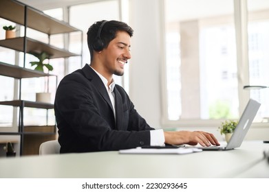Young man wearing wireless headset using a laptop computer for communication with customers or colleagues sitting at the desk in contemporary office. Maale support worker talks online. Side view - Shutterstock ID 2230293645