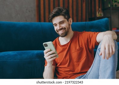 Young man wears red t-shirt hold in hand use mobile cell phone sit on blue sofa couch stay at home hotel flat rest relax spend free spare time in living room indoors grey wall. People lounge concept