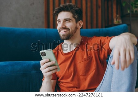 Young man wears red t-shirt hold use mobile cell phone look aside sit on blue sofa couch stay at home hotel flat rest relax spend free spare time in living room indoors grey wall People lounge concept