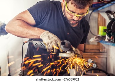 A Young Man Welder In A Black T-shirt, Goggles And Construction Gloves Processes Metal An Angle Grinder   In The Garage, In The Background A Lot Of Tools, Sparks Fly To The Side