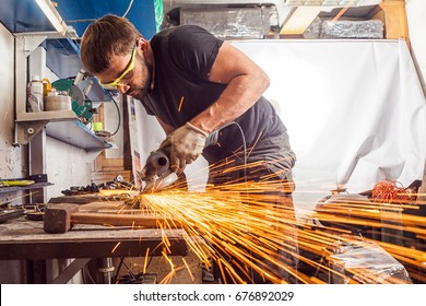 A young man welder in a black T-shirt, goggles and construction gloves grinder metal an angle grinder in the workshop, sparks fly to the side