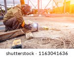 A young  man welder in brown uniform, welding mask and welders leathers, weld  metal  with a arc welding machine at the construction site, blue sparks fly to the sides