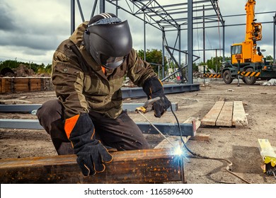 A young  man welder in brown uniform, welding mask and welders leathers, weld  metal  with a arc welding machine at the construction site, blue sparks fly to the sides - Shutterstock ID 1165896400