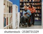 Young man in a wheelchair reading book with assistance of young woman in library filled with shelves of books. Shelves and stacks of books are prominently visible