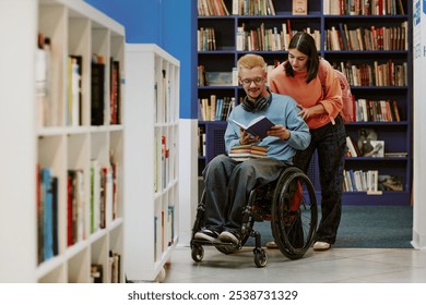 Young man in a wheelchair reading book with assistance of young woman in library filled with shelves of books. Shelves and stacks of books are prominently visible - Powered by Shutterstock