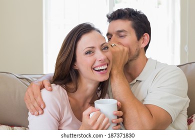 Young man whispering secret into a cheerful young womans ear in the living room at home - Powered by Shutterstock