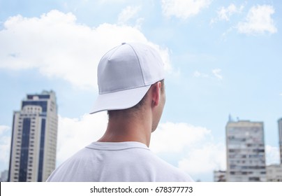 Young Man In A White Baseball Cap And White T-shirt On Background Of The City And Blue Sky With Clouds