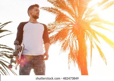 Young man in a white blank long sleeve t-shirt holding skateboard is looking aside while standing in the skate park on a warm sunny background. A skater is standing on the palm tree background.