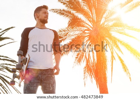 Young man in a white blank long sleeve t-shirt holding skateboard is looking aside while standing in the skate park on a warm sunny background. A skater is standing on the palm tree background.