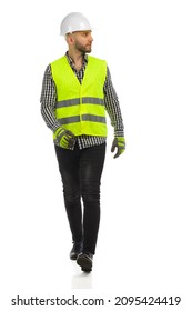 Young man in white hardhat, reflective waistcoat, lumberjack shirt and gloves is walking and looking to the side. Front view. Full  length studio shot isolated on white.