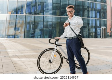 Young Man in a White Shirt Checks His Watch While Standing Next to His Bicycle Outside a Modern Glass Building During Daytime - Powered by Shutterstock