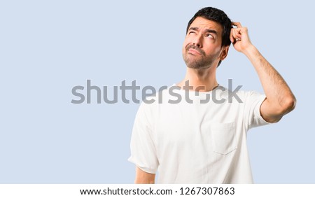 Young man with white shirt having doubts and with confuse face expression while scratching head on isolated blue background