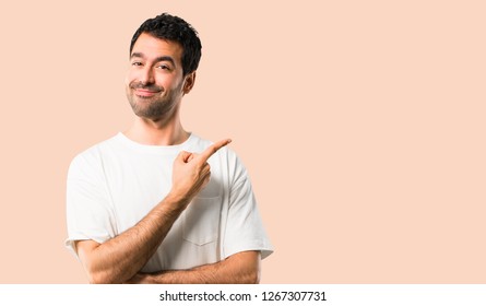Young man with white shirt pointing to the side with a finger to present a product or an idea while looking forward smiling on isolated ocher background