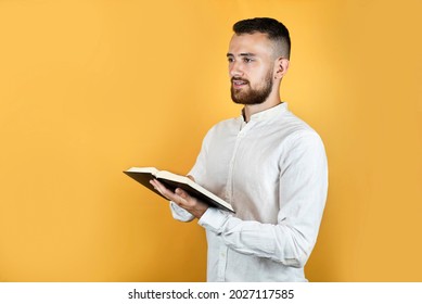 A Young Man In A White Shirt And Short Beard On A Yellow Background, Holding A Book In His Hands And Reading It And As If Saying Something, He Is Like A Preacher With A Bible In His Hands