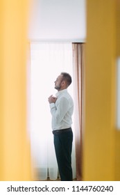 A young man in a white shirt stands at the window with yellow curtains - Shutterstock ID 1144760240