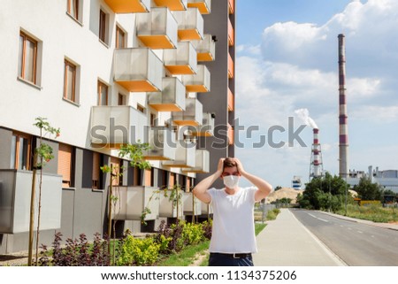 Young man in a white T-shirt and dark shorts posing near the entrance  to factory. Concept of a man in a mask near a new apartment building against the background of exhaust pipes.