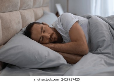 Young man in white t-shirt is in deep sleep, lying on his side on comfortable bedding, illustrating the importance of rest in serene bedroom setting - Powered by Shutterstock