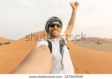 young man in white tshirt in desert, treveling in UAE on safari, wearing hat and backpack, exploring nature of sandy beauty. making selfie in the background of the sunset