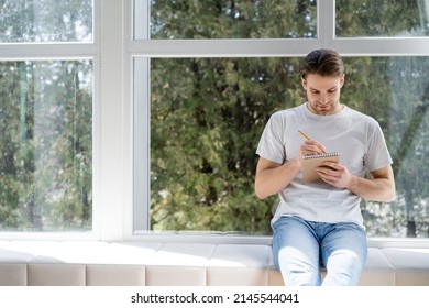 young man in white t-shirt and jeans sitting on windowsill and writing in diary