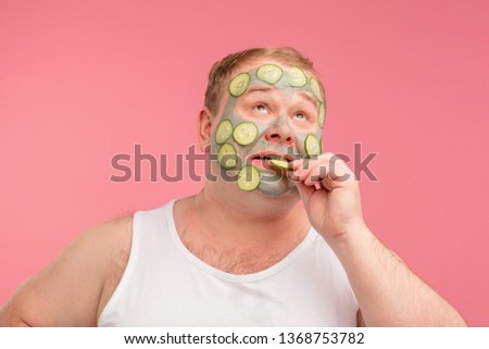 Young man in white undershirt having doubts and with confuse face expression while applying cucumber mask on his face, isolated pink background