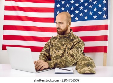 Young Man Who Serves In US Army Working On Laptop Computer. Serious Busy Soldier Who Is In USA Military Forces Using Modern Notebook PC While Sitting At Office Desk With American Flag In Background