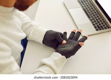 Young man who suffers from rheumatoid arthritis wearing pair of warm fingerless grey textile cotton spandex compression hand gloves for easing pain while working on notebook PC computer at office desk