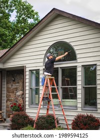 Young Man Window Stands On Ladder Holding Squeegee While Washing Outside Windows Of Midwestern House In Summer 