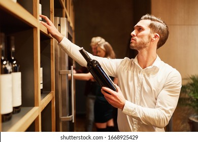 Young Man As A Wine Connoisseur Choosing Red Wine On The Shelf In The Wine Shop