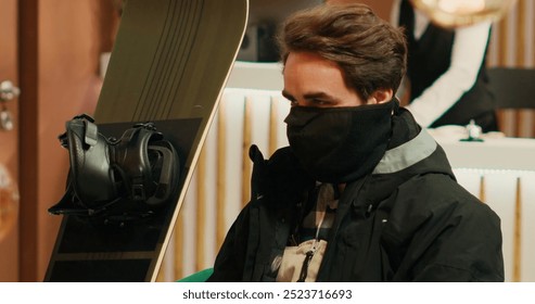 Young man in winter sport attire preparing to face the cold weather and go down snowy slopes with snowboard. Person sitting in lounge area at exclusive alpine resort waiting for check in. - Powered by Shutterstock