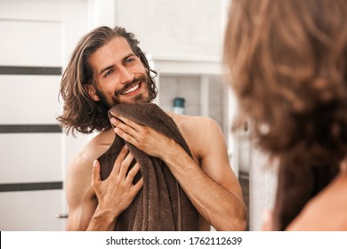 A young man wipes himself with a towel after a shower. A man smiles and looks in the mirror
