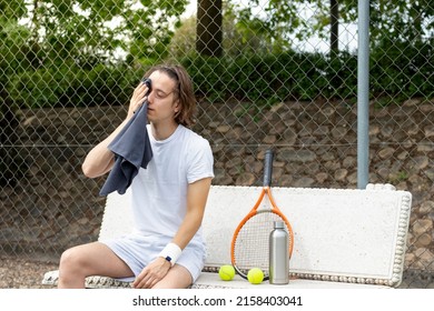 Young man wiping his sweat with a towel sitting on a bench after playing a game of tennis on a court - Powered by Shutterstock