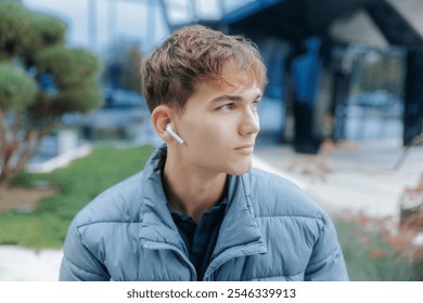 A young man with wireless earbuds stands in a city scene, dressed in a blue puffer jacket, focusing on something in the distance, representing modern youth and tech use. - Powered by Shutterstock