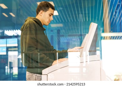 Young Man Withdrawing Money At An ATM In The City. Confused Business Man Using Credit Card Taking Money From An ATM Machine Inside A Branch Of Savings Bank. Side View Through Window.