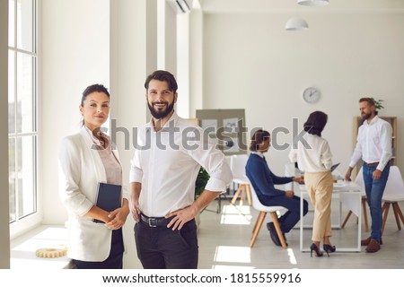 Young man and woman, 2 successful company co-founders, business partners or coworkers looking at camera. Confident female executive director and male manager standing in office after corporate meeting