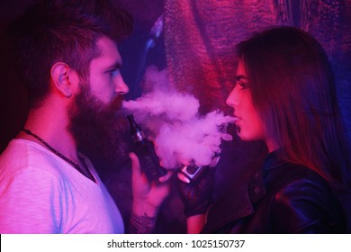 Young man and woman blowing smoke to join it in one cloud at black studio background. Relationship and vape addiction concept with copy space. Young sexy woman with bearded man are vaping together.