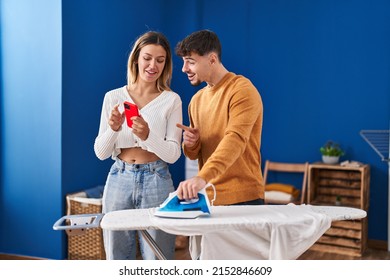 Young Man And Woman Couple Ironing Clothes Using Smartphone At Laundry Room