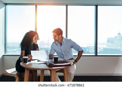 Young man and woman discussing work over a cup of coffee sitting in office. Businesswoman holding a paper in hand while taking to her business colleague.